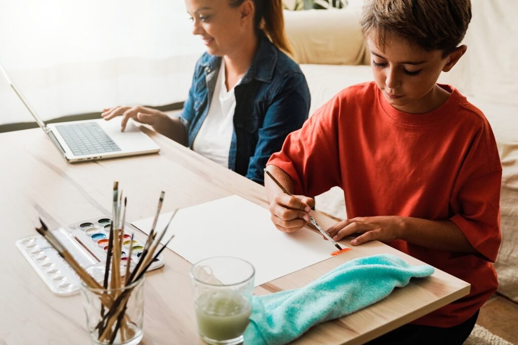 Happy Latin mother wor on computer child son is painting with watercolors - Home schooling